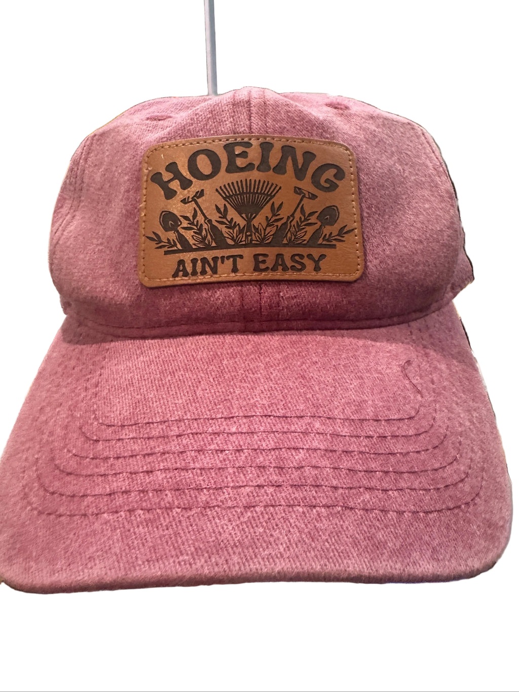 A dusty rose pink baseball cap with a brown leather patch on the front that reads 'HOEING AIN'T EASY' with a farming graphic. The cap is photographed in a cluttered workshop environment.