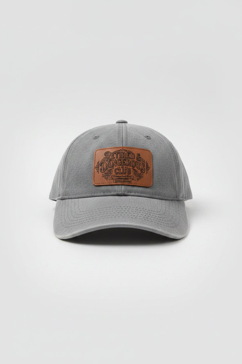 A heather gray baseball cap with a brown leather patch on the front that reads 'Retired & Dangerous Club'. The cap is photographed in a cluttered workshop or garage setting.