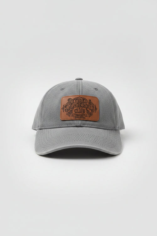 A heather gray baseball cap with a brown leather patch on the front that reads 'Retired & Dangerous Club'. The cap is photographed in a cluttered workshop or garage setting.
