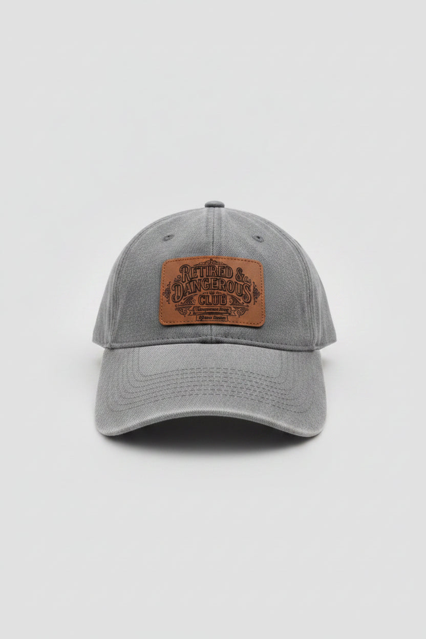 A gray washed baseball cap with a brown leather patch on the front that reads 'Bitter & Dangerous Club', photographed in a workshop or garage setting with tools and equipment in the background.