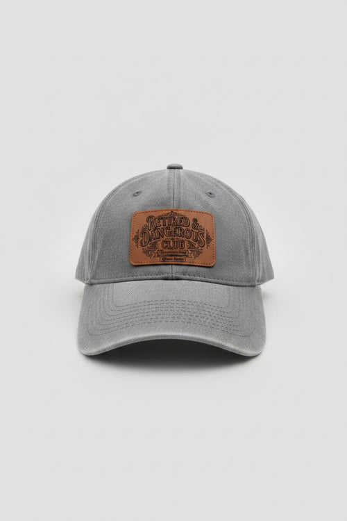 A gray washed baseball cap with a brown leather patch on the front that reads 'Bitter & Dangerous Club', photographed in a workshop or garage setting with tools and equipment in the background.